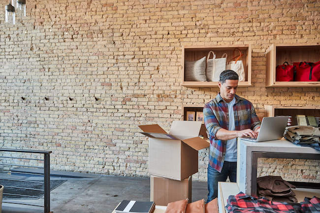 Man in a fashion store checking clothes stock on laptop while packing boxes