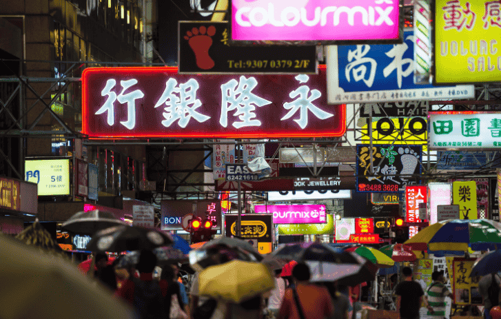 Rue commerçante très fréquentée vue de nuit, avec de nombreuses enseignes néon et des personnes tenant des parapluies.