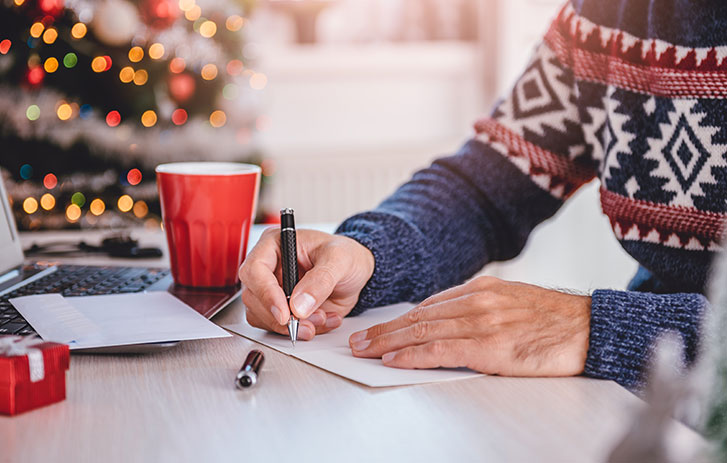 Man in holiday sweater writing card with Christmas tree in background 