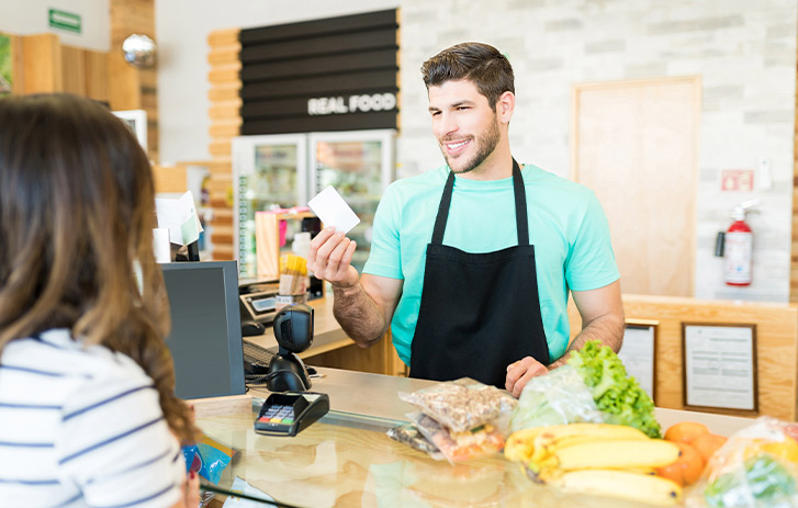 Smiling male owner giving reward card to customer at checkout counter in supermarket