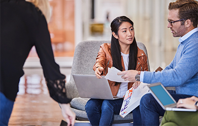 A man and a woman sit in an open office area, talking and working on their laptops.