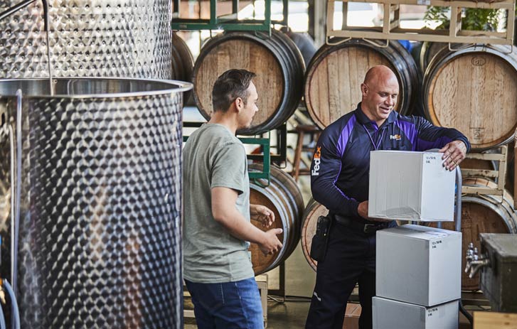 A photo of a FedEx worker stacking wine packages on a dolly while talking to a man near wine barrels