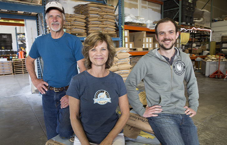 A photo of Dennis Meade, Diana Ambauen-Meade, and Byron Meade from Scratch and Peck sitting in a warehouse with feed bags in the background