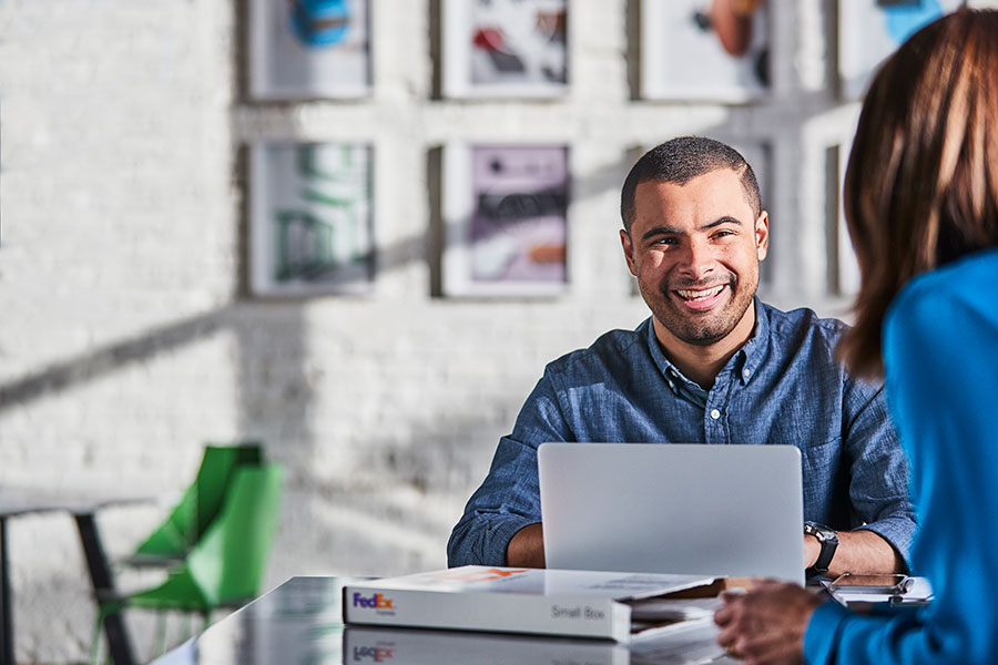 A man and a woman sitting at a table talking. The man is sitting in front of his laptop facing the camera, and the woman has her back to the camera. There is an open FedEx box in the middle of them.