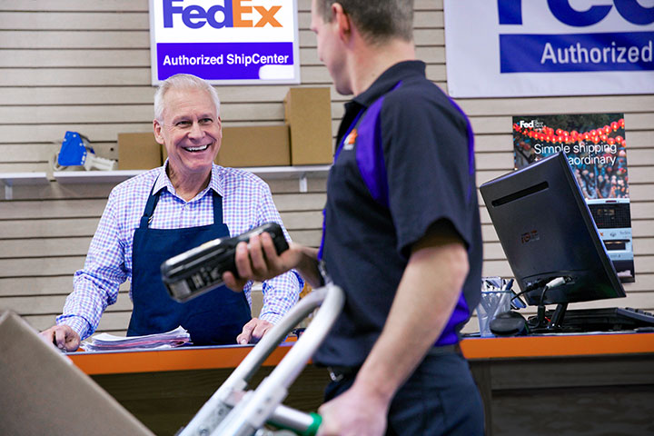 A FASC worker talking to a FedEx delivery driver with a dolly of packages inside a FASC store