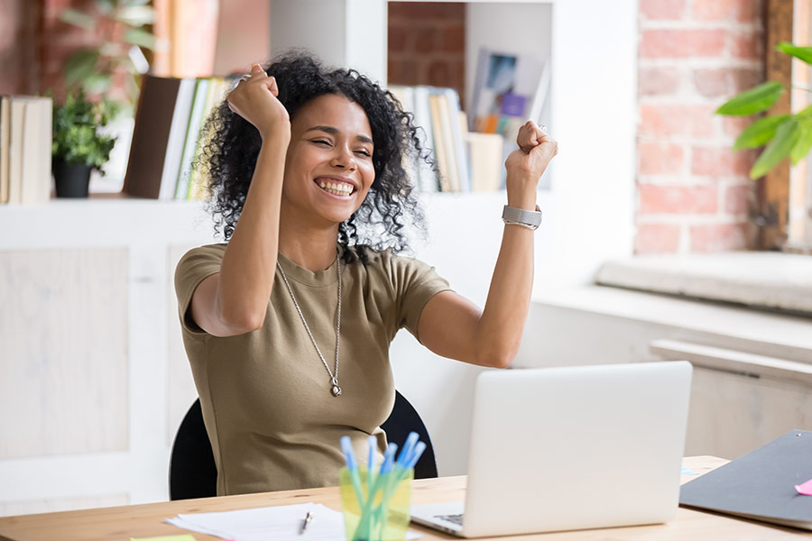 A woman smiling with her hands up cheerfully sitting as a desk in front of her laptop 