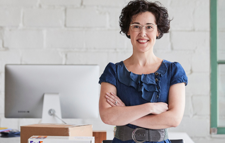 smiling woman standing in front of a work desk with her arms crossed and a FedEx box on the desk behind her